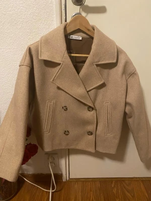 Beige ullkappa med bred krage - Stilren beige kappa från Zara i ullmix med bred krage och dubbelknäppning framtill. Kortare modell med raka ärmar och två fickor fram. Perfekt för dig som gillar klassisk men trendig look.