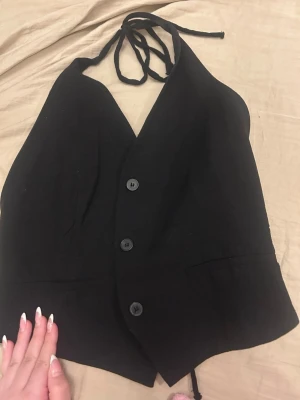 Svart halterneck korsettopp med knyt - Snygg svart halterneck korsettopp med knyt i nacken och midjan. Toppen har tre knappar framtill och en djup v-ringning. Perfekt för att skapa en edgy och trendig look. Materialet känns som bomull eller polyester.