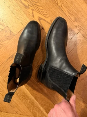 Svarta Chelsea boots från Loake - Klassiska svarta Chelsea boots från Loake i slätt läder. Skorna har elastiska sidopaneler, rundad tå och dragflikar både fram och bak med märkets logga. Perfekta för en stilren och tidlös look.