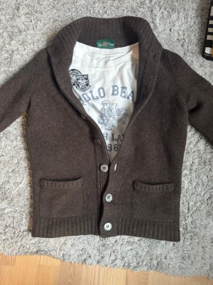 Alan Paine cardigan - Säljer en brun cardigan från Alan Paine i 100% lammull. Den har ett litet hål i armhålan, se bild, därav pris. Nypriset ligger på 2499kr. Perfekt för lager på lager. Storlek 46, sitter ungefär som en XS. Lappen vid nacken har gått upp pyttelite på ena sidan men inget som märks. Finns på sista bilden 