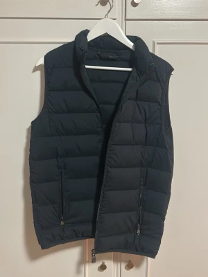 Svart dunväst med dragkedja  - Snygg svart dunväst med hög krage och två framfickor med dragkedja. Västen har quiltad design med breda kanaler och stängs med dragkedja framtill. Perfekt för lager-på-lager och ger en clean look till din outfit.