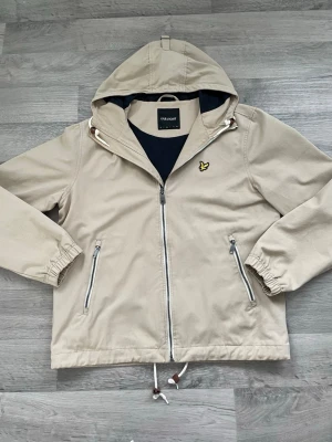 Beige vindjacka från Lyle & Scott - Stilren beige vindjacka från Lyle & Scott med huva, dragkedja framtill och två snedställda fickor med dragkedja. Jackan har elastiska muddar vid ärmsluten, justerbar dragsko i nederkant och svart innerfoder. Ikonisk gul logga på bröstet.