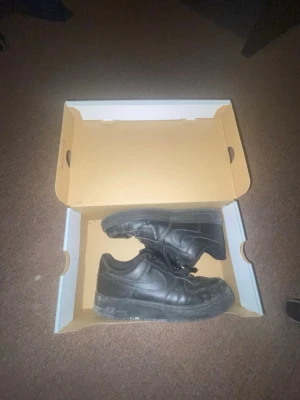 Svarta Nike Air Force 1 sneakers - Klassiska helsvarta Nike Air Force 1 sneakers i läder med låg profil och ikonisk swoosh på sidan. Skorna har tjock sula och snörning framtill. Perfekta för dig som gillar streetwear och tidlös stil.