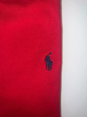 Röd t-shirt från Ralph Lauren - Klassisk röd t-shirt från Ralph Lauren med den ikoniska mörkblå polospelaren broderad på bröstet. T-shirten har rund halsringning och är tillverkad i mjuk bomull för en skön känsla. Perfekt till jeans eller shorts för en clean look. 