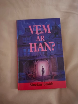 Vem är han? av Sinclair Smith - En mystisk och spännande bok med titeln 'Vem är han?' av Sinclair Smith. Omslaget är blått och rött med en siluett av en person framför ett gammalt hus. Boken är inbunden och riktar sig till dig som gillar thrillers och mysterier.