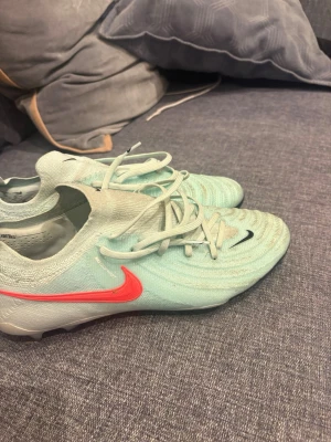 Nike ljusblå fotbollsskor med orange swoosh - Säljer ett par stilrena ljusblå Nike fotbollsskor med orange swoosh på sidan och svarta detaljer. Skorna har snörning och är tillverkade i ett syntetmaterial som ger bra grepp på planen. Perfekta för dig som vill sticka ut på matchen! Skorna är endast använda 1 träning på konstgräs. Hör av dig vid intresse!