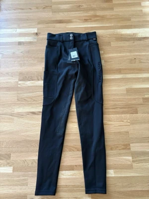 Svarta ridleggings från Kingsland - Snygga svarta ridleggings från Kingsland med bälteshällor och två knappar fram. Helskoda och praktiska fickor på sidorna. Perfekta för dig som vill ha både stil och funktion i stallet. OBS prislappen kvar aldrig använda 