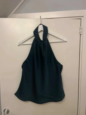 Mörkgrön halterneck linnetopp - Snygg mörkgrön halterneck topp med bred krage som knyts i nacken. Toppen är ärmlös och har en lös passform, perfekt för en trendig och avslappnad stil. Tillverkad i ett mjukt och lätt material som känns skönt mot huden.