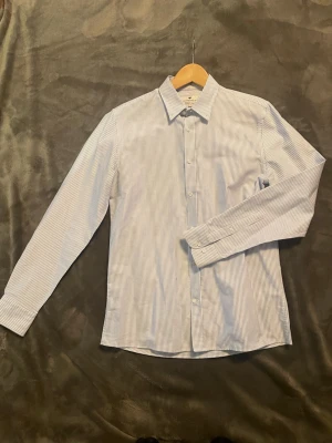 Randig skjorta från Berns Edition - Säljer en klassisk skjorta från Berns Edition i vitt och ljusblått med tunna ränder. Skjortan har lång ärm, knappar framtill och en snygg krage. Perfekt för dig som gillar stilrena och tidlösa plagg. Tveka inte att höra av dig vid funderingar