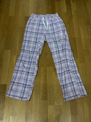 Lila rutiga pyjamasbyxor - Mysiga pyjamasbyxor i lila och vitt rutigt mönster med snören i midjan. Byxorna har raka ben men lite utsvängda nedtill och resår i midjan för extra komfort. Perfekta för chill hemma eller när du vill vara bekväm.