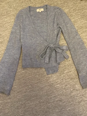 Ljusblå knytkofta från Gina Tricot - Supersöt ljusblå stickad omlottkofta från Gina Tricot i mjuk ullblandning. Koftan har långa ärmar, ribbade muddar och knyts i sidan med ett brett band. Perfekt för lager på lager och ger en chill men ändå snygg look.