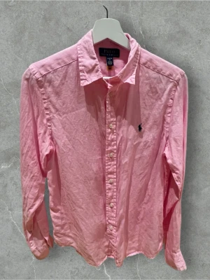 Rosa skjorta från Polo Ralph Lauren - Klassisk rosa skjorta från Polo Ralph Lauren med lång ärm och knappar framtill. Skjortan har den ikoniska broderade loggan på bröstet och är tillverkad i bomull. Perfekt för en clean och stilren look.