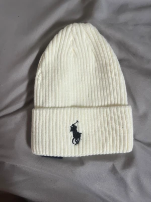Vit ribbstickad mössa från Polo Ralph Lauren - Snygg vit ribbstickad mössa från Polo Ralph Lauren med uppvikt kant och klassiska loggan broderad framtill i svart. Tillverkad i mjukt material som håller dig varm och stilren under kalla dagar.