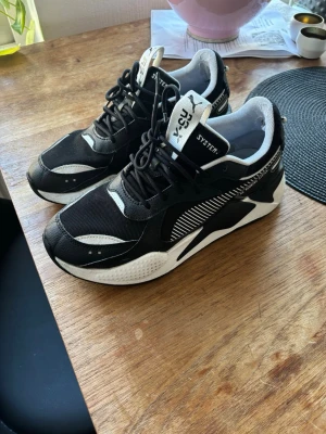 Svarta Puma RS-X sneakers - Säljer ett par svarta och vita Puma RS-X sneakers med coola detaljer och meshpaneler. Skorna har en chunky sula, vita och svarta kontraster samt RS Running System-tryck på sidan. Perfekta för dig som gillar streetwear och sportig stil.