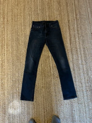 Mörkblå smala jeans från Nudie Jeans - Säljer ett par mörkblå jeans från Nudie Jeans med smal passform och klassisk femficksdesign. Jeansen har ett litet hål i skrevet och är tillverkade i denim med en lätt stretch för extra komfort. Perfekta för dig som gillar en stilren och modern look.