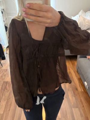 Brun transparent blus med knytband - Snygg brun blus i transparent material med långa ärmar och v-ringning. Blusen har volangdetaljer och knytband framtill som ger en avslappnad och trendig look. Perfekt att styla med jeans eller kjol för en cool vibe.