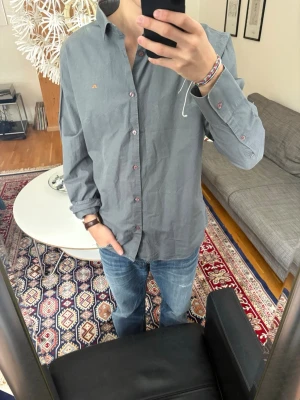 Grå skjorta från J.Lindeberg - Stilren grå skjorta från J.Lindeberg med klassisk krage och knappar i vinrött. Liten orange logga broderad på bröstet. Vintage.