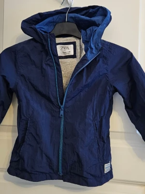 Mörkblå vindjacka från Zara str 98 - Mörkblå jacka från Zara med huva och dragkedja i matchande blå färg. Jackan har två sidofickor .
