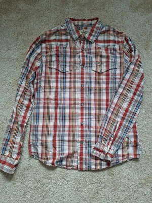 Rutig skjorta från Wrangler, slim fit - Säljer en rutig långärmad skjorta från Wrangler i slim fit. Skjortan har knappar framtill, två bröstfickor med lock och är i flera färger som röd, blå, beige och vit. Perfekt för dig som gillar klassisk stil med modern passform.