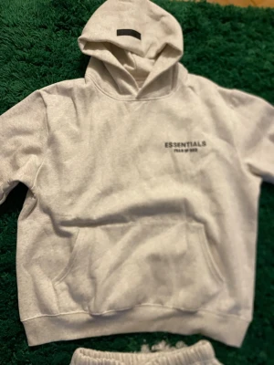 Grå hoodie från Essentials - Grå hoodie från Essentials med huva och magficka. Tillverkad i mjukt material och har ett diskret tryck på bröstet. Perfekt för en avslappnad och trendig stil. 