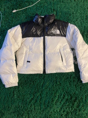 Vit och svart dunjacka The North Face - Säljer en snygg dunjacka från The North Face i vit och svart. Jackan har hög krage, dragkedja framtill och två stora fickor med dragkedja. Klassisk boxig passform och logga på bröstet. Perfekt för kalla vinterdagar och riktigt trendig stil.