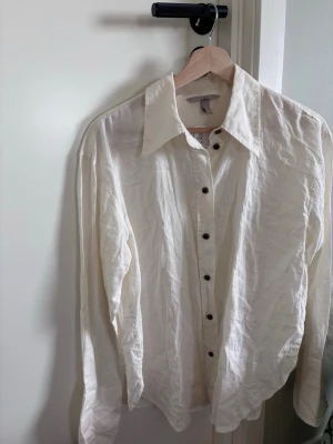 Vit linneskjorta med knappar - Klassisk vit skjorta i linne med lång ärm och bruna knappar. Skjortan har en avslappnad passform och stor krage, perfekt för en stilren och enkel look. Passar till många olika outfits och är lätt att matcha.