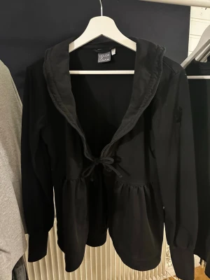 Svart volangtröja från House of Lola - Svart långärmad tröja från House of Lola med volang kring midjan och stor rynkad krage🤍 Tröjan har knytning framtill och är av mjukt och stretchigt material. Bara använd ett fåtal gånger🤍