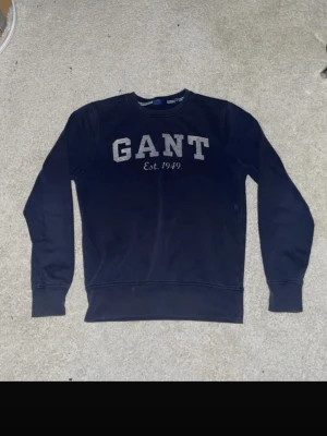 Mörkblå sweatshirt från GANT - Mörkblå sweatshirt från GANT med stor logga och texten 'Est. 1949' på bröstet. Klassisk rund halsringning och ribbade muddar vid ärmslut och nederkant. Tillverkad i mjukt bomullsmaterial som är skönt att bära.