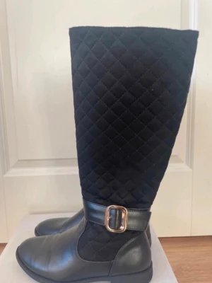 Svarta quiltade stövlar med spänne - Snygga svarta stövlar med quiltat tygskaft och detaljer i konstläder. Stövlarna har ett dekorativt guldfärgat spänne vid ankeln och rundad tå. Perfekta för dig som vill ha en trendig och bekväm look till hösten eller vintern.