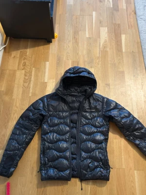 Svart Peak performance helium glacier - Snygg svart pufferjacka med huva och vågigt quiltat mönster. Jackan har dragkedja framtill och två sidofickor med dragkedja. Materialet är glansigt och ger en modern look. Perfekt för kalla dagar när du vill hålla dig varm och stilren. Köpt för 3200kr