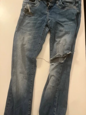 Blå slitna jeans från LTB  - Säljer ett par blå jeans från LTB med slitningar och hål på låret och vid fickan. Modellen har klassisk femficksdesign, dragkedja och knapp. Jeansen har en tvättad look och är perfekta för en avslappnad stil.