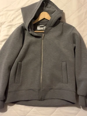 Grå hoodie med dragkedja från Amisu - En stilren grå hoodie från Amisu med dragkedja framtill och två fickor. Tröjan har en klassisk huva och är gjord i mjukt sweatshirtmaterial. Perfekt för en avslappnad look och enkel att matcha med andra plagg.