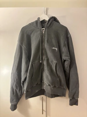 Grå zip hoodie från Stüssy - Säljer en grå zip hoodie från Stüssy med vit logga på bröstet. Hoodien har dragkedja, två fickor framtill och justerbar huva med snören. Tillverkad i mjukt bomullsmaterial och har en avslappnad passform. Perfekt för en chill streetwear-look. Den är i storlek M och är tts