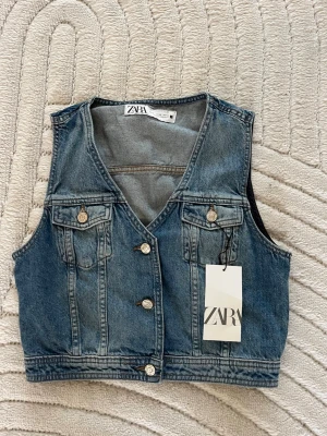 Blå jeansväst från Zara - Snygg blå jeansväst från Zara med klassisk v-ringning och knappar framtill. Västen har två bröstfickor med lock och silverfärgade detaljer. Tillverkad i denim för en trendig och avslappnad look.