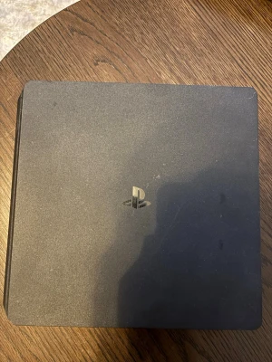 PlayStation 4 Slim svart konsol - Säljer en svart PlayStation 4 Slim från Sony. Konsolen har en stilren, rektangulär design med rundade hörn och PlayStation-loggan på ovansidan. Kablar, kontroll och laddare får man. Jätte bra skick fungerar perfekt