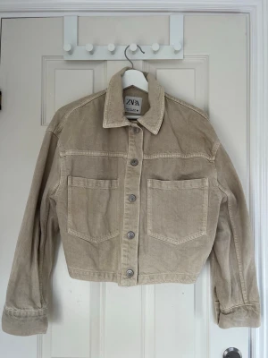 Beige jeansjacka från Zara - Snygg beige jeansjacka från Zara med klassisk krage, raka axlar och två stora bröstfickor. Jackan har knäppning framtill med metallknappar och kan dras åt i midjan. Perfekt för en avslappnad och trendig look.