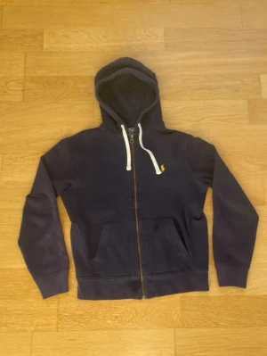 Mörkblå hoodie från Polo Ralph Lauren - Mörkblå hoodie med dragkedja från Polo Ralph Lauren. Klassisk design med vit dragsko i huvan, två fickor fram och liten gul broderad logga på bröstet. Tillverkad i mjukt och skönt material, perfekt för chill dagar.