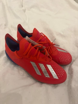 Röda Adidas fotbollsskor med silverdetaljer - Snygga fotbollsskor från Adidas i rött med silvriga ränder på sidan och blå detaljer vid hälen. Skorna har snörning, låg profil och är tillverkade i syntetmaterial för bra grepp och kontroll på planen. Perfekta för dig som vill sticka ut på fotbollsplanen. HELT NYA
