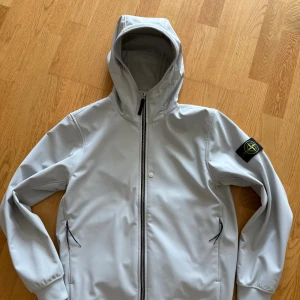Ljusgrå softshelljacka från Stone Island - Storlek M. Ljusgrå softshelljacka från Stone Island med huva, svarta dragkedjor och ikonisk patch på ärmen. Jackan är tillverkad i stretchig polyester med fleecefoder och har två fickor med dragkedja. Perfekt för dig som gillar stilren och funktionell streetwear. Knappt använd då den jag köpte fel storlek. 