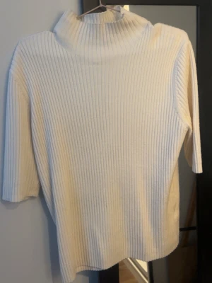 Vit ribbstickad polotröja - Säljer en stilren vit ribbstickad polotröja med halvlång ärm och hög krage. Tröjan har en rak passform och är gjord i ett mjukt, stretchigt material som känns skönt mot huden. Perfekt att matcha med jeans eller kjol för en clean look. 