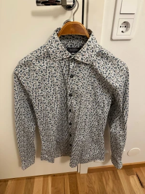 Blommig skjorta från Dressmann - Snygg långärmad skjorta från Dressmann med småblommigt mönster i blått och brunt på vit botten. Skjortan har klassisk krage och knäppning framtill. Perfekt för dig som gillar stilrena men ändå lekfulla plagg.