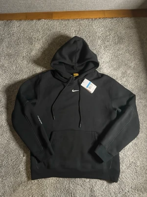 Svart Nike hoodie med logga - Snygg svart hoodie från Nike med klassisk vit Swoosh-logga på bröstet. Hoodien har justerbar dragsko i huvan och en stor magficka framtill. Tillverkad i mjukt material som känns skönt mot huden. Perfekt för en avslappnad och sportig stil.