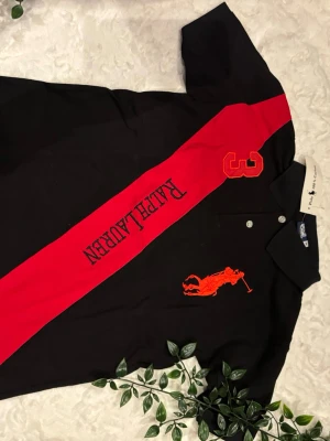 Svart och röd skjorta från Ralph Lauren - Svart skjorta från Ralph Lauren med röd diagonal rand, stor broderad polospelare och siffran 3 på bröstet. Klassisk krage och knappar framtill. Perfekt för dig som gillar sportig och stilren design.