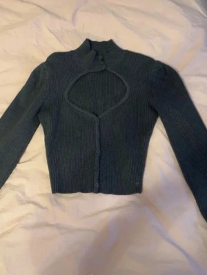 Mörkgrå ribbad cardigan med cut-out - Trendig mörkgrå ribbad cardigan med lång ärm och unik cut-out detalj vid bröstet. Kortare modell med knappar framtill och hög krage. Perfekt för dig som vill sticka ut med din stil.