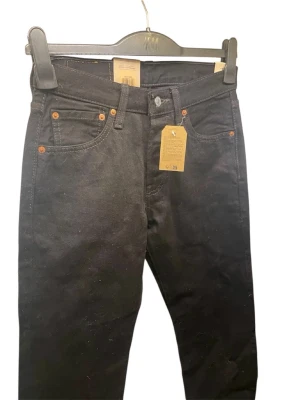 Svarta Levi's 501 jeans - Klassiska svarta Levi's 501 jeans med raka ben och femficksdesign. Jeansen har knäppning framtill, orangea nitar och är tillverkade i slitstark bomullsdenim. Perfekta för dig som gillar en tidlös och clean look.