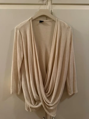 Beige omlottopp från Gina Tricot - Snygg beige omlottopp från Gina Tricot med djup v-ringning och draperad front. Toppen har trekvartsärm och är tillverkad i ett mjukt, tunt material som känns skönt mot huden. Perfekt för dig som gillar en avslappnad men trendig stil.