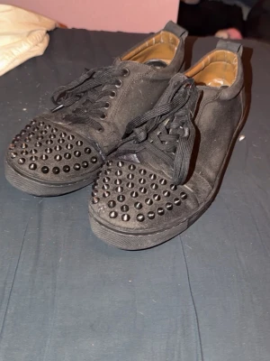  Christian Louboutin skor - Snygga svarta sneakers från Christian Louboutin med svarta nitar på tån och klassisk röd sula. KOLLA ALLA BILDER! Skorna har snörning och är tillverkade i textil och läder. Perfekta för dig som vill sticka ut med en edgy och lyxig stil. 1 nit av. Och andra defekter som visas noga på alla bilder! Därav priset. Lite smutsiga under, går nog bort vid tvättning!💓 skriv för frågor eller så! Öppen för prisförslag också!