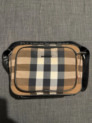 Burberry axelväska i beige rutigt - Säljer en snygg axelväska från Burberry med klassiskt beige, svart och vitt rutmönster. Väskan har en svart axelrem med Burberry-logga och en praktisk ytterficka med dragkedja. Tillverkad i tåligt canvasmaterial med detaljer i svart.