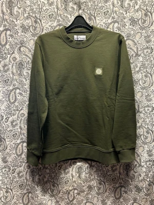 Olivgrön sweatshirt från Stone Island - Olivgrön sweatshirt från Stone Island med rund halsringning och liten broderad logotyp på bröstet. Tröjan har ribbade muddar vid ärmslut och nederkant, och är tillverkad i mjukt material. Perfekt för en avslappnad och stilren look. Finns i flera storlekar, skriv önskad storlek i samband lagd order. 