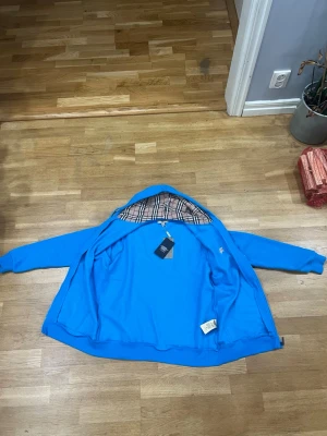 Blå fleece från Burberry - Snygg blå vindjacka från Burberry med klassiskt rutigt foder på insidan av huvan. Jackan är tillverkad i 65% bomull och 35% polyester, har dragkedja och ribbade muddar vid ärmsluten. Perfekt för dig som vill ha en stilren och ikonisk look.